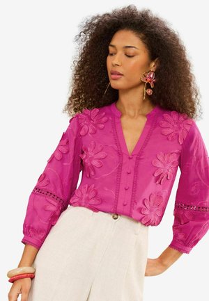 Woman with curly hair wearing bright pink floral-embroidered blouse, beige high-waist pants, large floral earrings, and bangles on wrist.