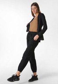 Černá lehká bunda přes hnědý top, spárovaná s černými užšími kalhotami a černými sportovními botami. Hladká textilie a volný design.