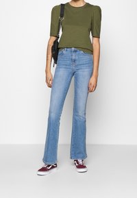 Oliven grøn pufærmet top, lyseblå flagrende jeans og maroon Vans sneakers. Sort crossbody taske og synlige tatoveringer på underarmen.