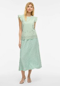 Mátově zelený top s řasenými rukávy, okraj s výřezovými detaily. Vyhovující strukturovaná sukně, po kotníky, ve stejné mátové barvě. Stříbrné sandály.