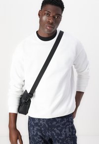 Vit crewneck-tröja med ribbade ärmslut, i kombination med mörka mönstrade byxor. En liten svart axelväska bärs över kroppen.