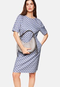 Blau gemustertes Kleid mit kurzen Ärmeln und Taschen, kombiniert mit einer grauen, geschwungenen Handtasche mit langen Fransen.