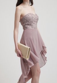 Šaty bez ramínek s krajkovým korzetem ve světle mauvové barvě a volnou šifonovou sukní. Mají asymetrický lem a béžovou kabelku se cvočky.