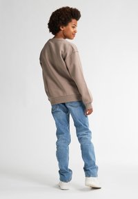 Felpa oversize marrone con polsini a coste, abbinata a jeans dritti blu chiaro. Il modello indossa sneakers bianche e si trova in posizione leggermente decentrata.