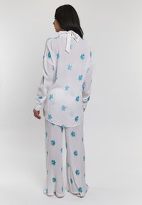 Witte outfit met een long-sleeve top en wijde broek, beide versierd met turquoise bladeren en sterrenpatronen. Rugstrikdetail op de top.