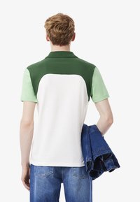 Paita polo dengan desain blok warna hijau dan hijau muda. Terbuat dari kain yang lembut, dilengkapi dengan kerah. Model memegang jaket denim biru.