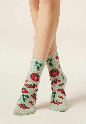 Gambe con calzini alla caviglia verde chiaro decorati con pomodori rossi e foglie verdi su uno sfondo uniforme.
