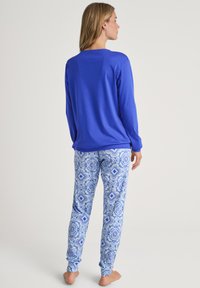 Maglietta blu a maniche lunghe abbinata a pantaloni da pigiama blu e bianchi a motivi floreali. Il modello è in piedi a piedi nudi, rivolto di spalle.