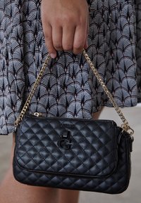 Hånd holder en sort quiltet læder GUESS håndtaske med guld kædetræ, mod en mønstret nederdel baggrund.