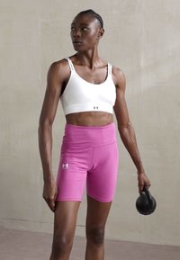 Bijela sportska grudnjak i ružičaste tajice koje se dobro prilagođavaju tijelu s detaljem logotipa. Model stoji držeći crnu kettlebell, prikazujući atletsku odjeću.
