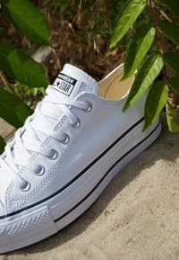 Zapatilla de lona blanca con acentos negros, puntera redonda y diseño de cordones. Cuenta con una suela de goma y un dibujo texturizado para mayor agarre.