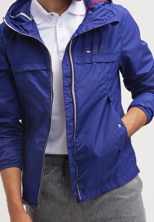 Homme portant une veste coupe-vent bleue zippée par-dessus un polo blanc avec un pantalon gris, une main dans la poche de la veste.