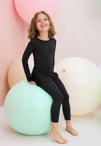 Vaikas, sėdintis ant šviesiai žalios treniruoklių kamuolio, dėvi juodą ilgarankovį marškinėlį ir tamprės. Fone matomi švelnių pastelinių spalvų balionai.