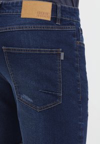 Sötétkék farmernadrág hátizsebbel, barna bőr címkével, rajta a "DENIM JOURNEY" felirattal és látható varrási részletekkel. Egységes textúra.
