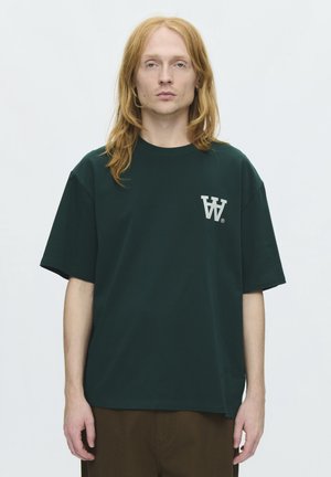 Jeune adulte aux longs cheveux roux portant un t-shirt oversize vert foncé avec un logo double W blanc sur la poitrine, debout devant un fond uni.