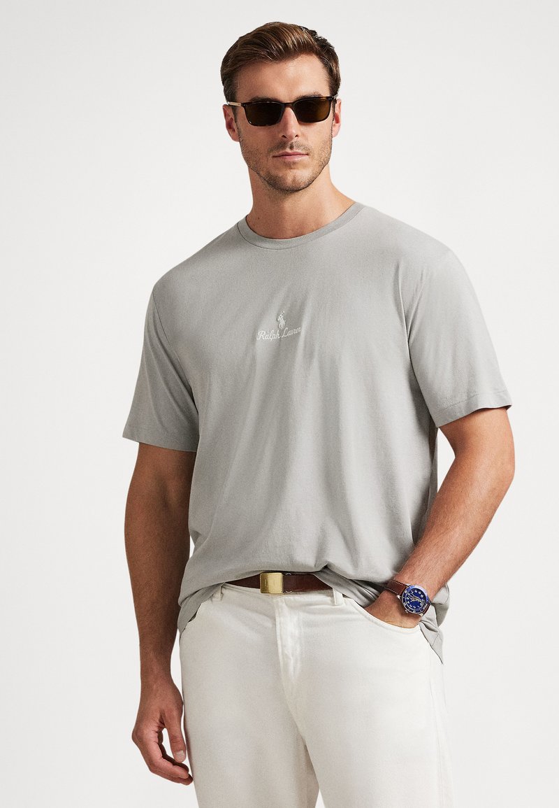 Lysegrå bomulls-T-skjorte med rund hals, med en liten, brodert logo. Brukes sammen med hvite bukser og et brunt belte, aksentrert med solbriller.