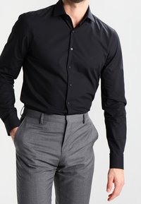 Svart, långärmad skjorta med knäppning och krage. Bärs med grå, skräddarsydda byxor som har en slät tygfinish.