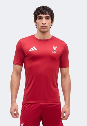 Mand iført rød Liverpool FC fodboldtrøje med Adidas-logo på brystet, står mod en ensartet lys baggrund.