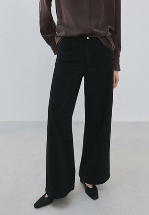 Jeans negros de pierna ancha con cintura alta, con diseño clásico de cinco bolsillos y cierre con botón plateado, combinados con una blusa oscura y zapatos negros.