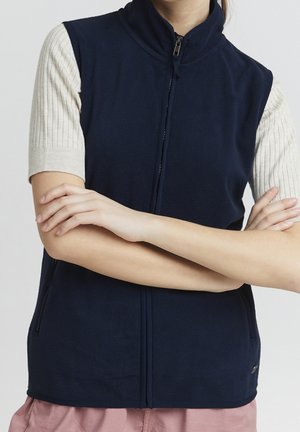 Henkilö, joka käyttää laivastonsinistä hihnatonta vetoketjuvaa liiviä kermanvärisenä ribbineulottua lyhythihaista puseron päällä, kädet ristissä vartalon edessä.