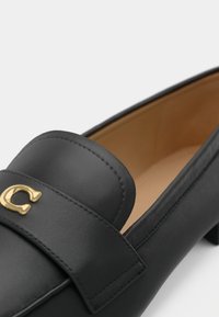 Black leather loafer with a rounded toe, featuring a gold-accented logo, stitched detailing, and a smooth interior lining.