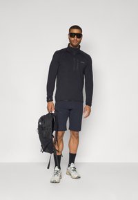 Zwarte ritsfleece, zwarte shorts, zwarte rugzak, grijze sneakers met zwarte accenten. Model draagt oversized zonnebril. Minimale achtergrond.