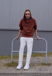 Brun Nike sweatshirt med svart logotyp, vita Nike sweatpants och vita sneakers. Personen lutar sig mot ett metallräcke mot en grå vägg.