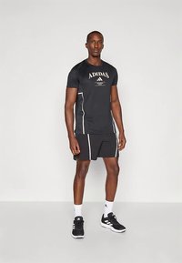 Must Adidas treeningpluus valgete detailide ja logoga, kombineeritud mustade lühikestega. Modell kannab musti spordijalatseid ja valgeid sokke.