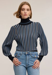 Pruhovaný svetr s vysokým límcem, s modr�ými, šedými a černými vertikálními pruhy; měkká textura a nabírané rukávy. V kombinaci se světle modrými džíny.