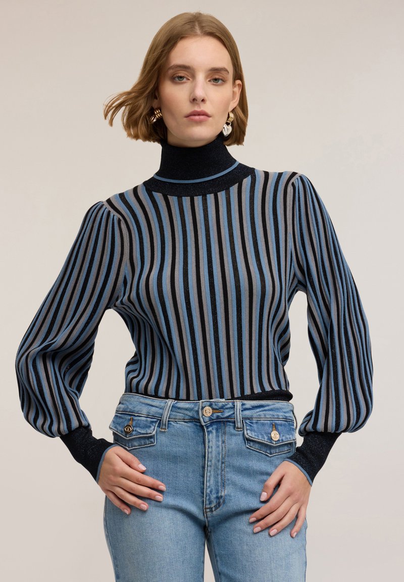 Pruhovaný svetr s vysokým límcem, s modrými, šedými a černými vertikálními pruhy; měkká textura a nabírané rukávy. V kombinaci se světle modrými džíny.