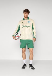 Conjunto de fútbol verde y crema con mangas largas, un collar y detalles del logo. El modelo sostiene un balón de fútbol, vistiendo shorts verdes y zapatos negros.