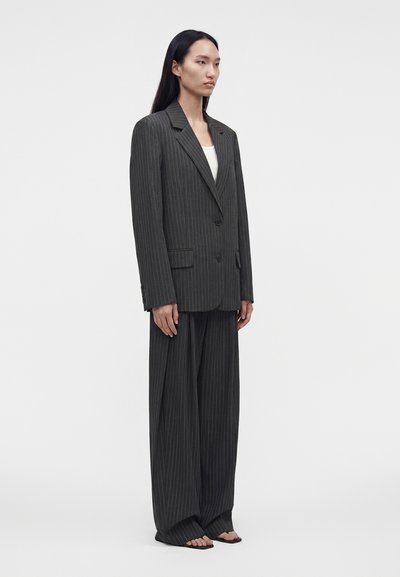 Femme debout dans un costume oversize à rayures foncées avec pantalon large, haut blanc, sandales et cheveux raides sur fond blanc uni.