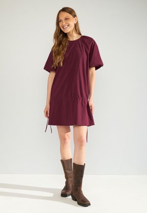 Donna sorridente con capelli lunghi castani, indossa un vestito largo bordeaux e stivali marroni fino al ginocchio, in piedi davanti a uno sfondo chiaro e uniforme.