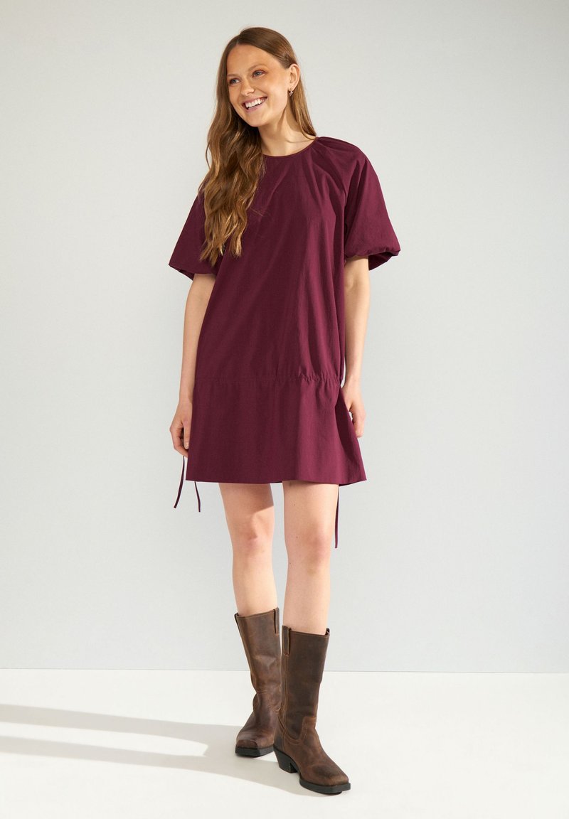 Femme souriante aux longs cheveux bruns, portant une robe ample couleur bordeaux et des bottes marron montant jusqu’aux genoux, debout devant un fond clair uni.