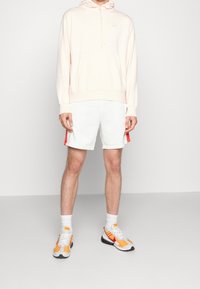 Ljusrosa hoodie med snörning, vita shorts med röda sidostråk och sneakers med orangea accenter. Vita strumpor avslutar looken.