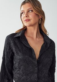 Zwarte shirt gemaakt van gestructureerde, gekreukelde stof met een kraag en een knoopsluiting. Heeft een diepe V-hals en lange mouwen.