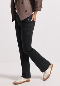 Jean noir évasé en denim, offrant un ajustement serré aux cuisses et une coupe décontractée aux chevilles. Assorti à des baskets beiges et blanches.