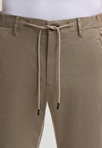 Beige broek van gestructureerde stof met een koord in de taille. Voorzien van een voorzak en contrasterende zwarte punten op het koord.
