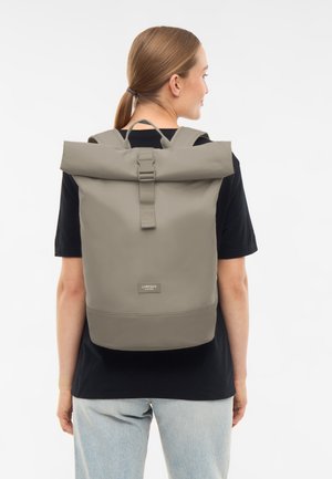 Donna con capelli castano chiaro raccolti, che indossa una maglietta nera e jeans, che porta uno zaino beige con chiusura a rotolo con l'etichetta "Larkson".