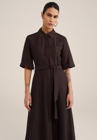 Rochie cămașă de lungime până la genunchi, de culoare maro, fabricată dintr-un material ușor. Prezintă mâneci scurte, două buzunare pe piept, un cordon în talie și o textură netedă.