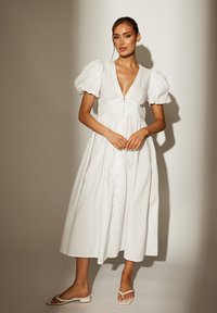 Weißes, knielanges Kleid mit Puffärmeln, tiefem V-Ausschnitt und durchgehender Knopfleiste. Hergestellt aus einem leichten Stoff; glatte Textur und fließendes Design.