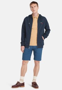Felpa con zip blu navy, con tasche a marsupio, indossata sopra una polo beige e pantaloni shorts blu. Scarpe da barca marroni con calze crema. Outfit casual.