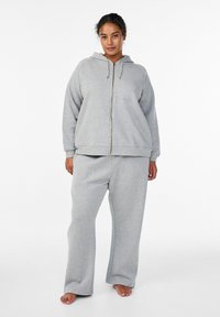 Felpa grigia con zip e cappuccio regolabile, abbinata a pantaloni della tuta ampi coordinati. Tessuto morbido, polsini e fascia in vita a coste.