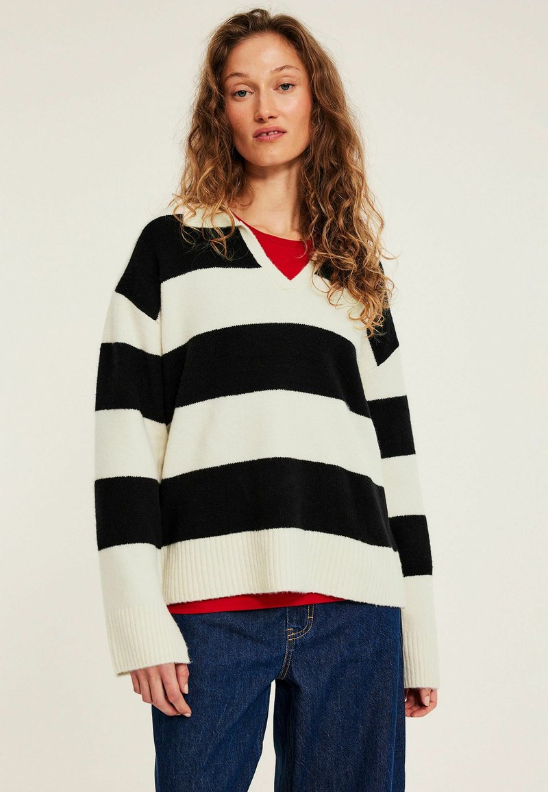 Sort- og hvidstribet V-hals sweater med ribkant forneden og ved ærmerne, lagdelt over en rød top. Løs pasform med lange ærmer. Blød tekstur.