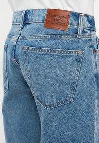 A kékt denim farmernadrág magas derekú, bőr márkacímkével, két hátsó zsebbel díszvarrással, és enyhén kopott hatásúra van elkészítve.