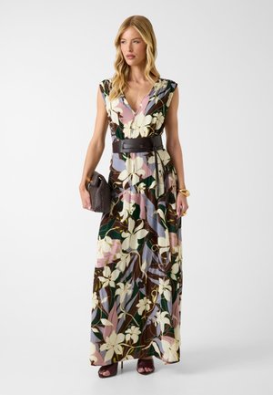 Kvinde iført en blomstret ærmeløs maxikjole med sort bælte, der holder en tekstureret clutch og iført brune sandaler med hæl.