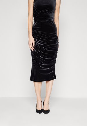 Robe mi-longue sans manches en velours noir avec des détails froncés, portée avec des escarpins noirs à bout pointu, vue des épaules aux pieds.