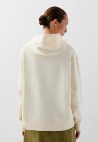 Kvinde med hår i en knold iført en off-white hættetrøje og en olivengrøn nederdel, vendt væk mod en enkel hvid baggrund.