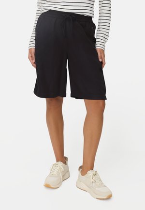 Person iført sorte shorts med snøre, stribet langærmet skjorte og beige sneakers står mod en hvid baggrund.