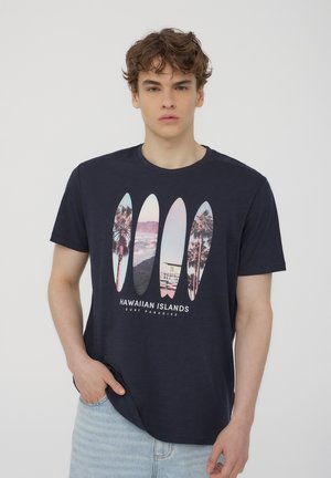 T-shirt in cotone blu navy con una grafica di cinque tavole da surf, che include palme e immagini costiere, con il testo "ISOLE HAWAIANE" sotto.